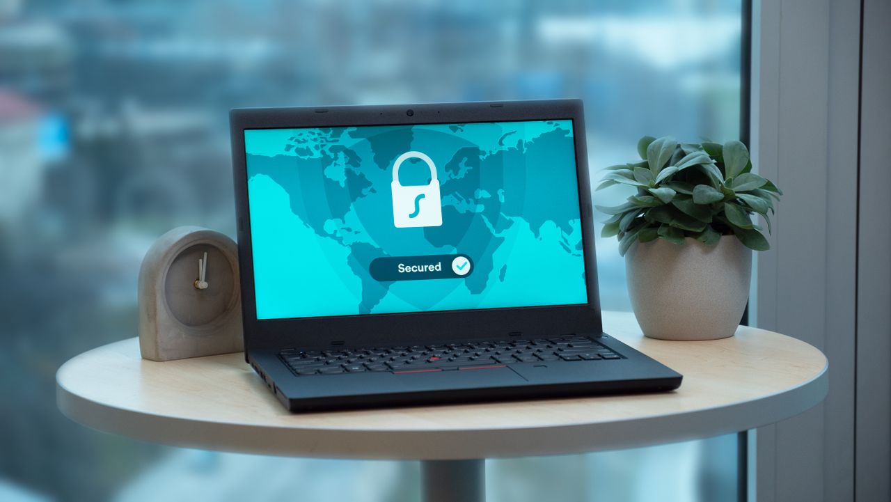 Home Laptop displaying a security lock icon on a table with a potted plant and clock.